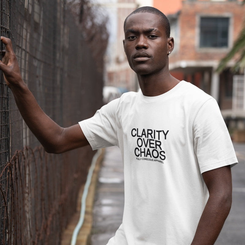 A man wearing a white t-shirt with the black printed text 'CLARITY OVER CHAOS' and 'BUILD A HARROGATESTATION' underneath, leaning against a wire fence in an outdoor urban setting.