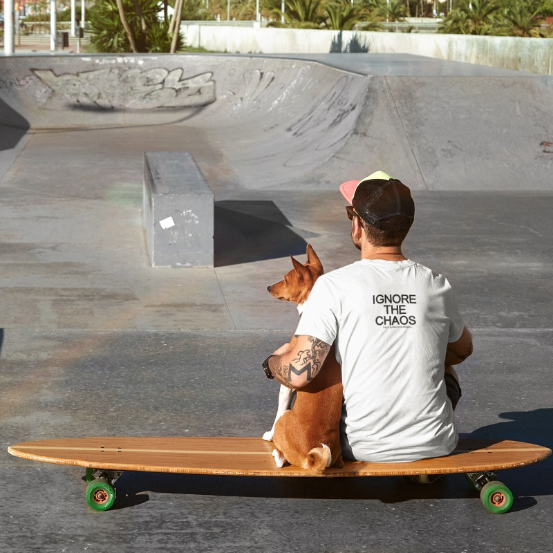 Man at a skatepark sitting on a longboard with his dog, wearing white ‘IGNORE THE CHAOS™’ tee from Fully Conscious Apparel, back view, modern streetwear motivational shirt promoting clarity and calm mindset.