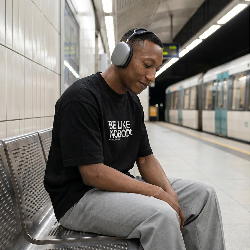 Man in a black BE LIKE NOBODY t-shirt by Fully Conscious Apparel sitting on a subway station bench